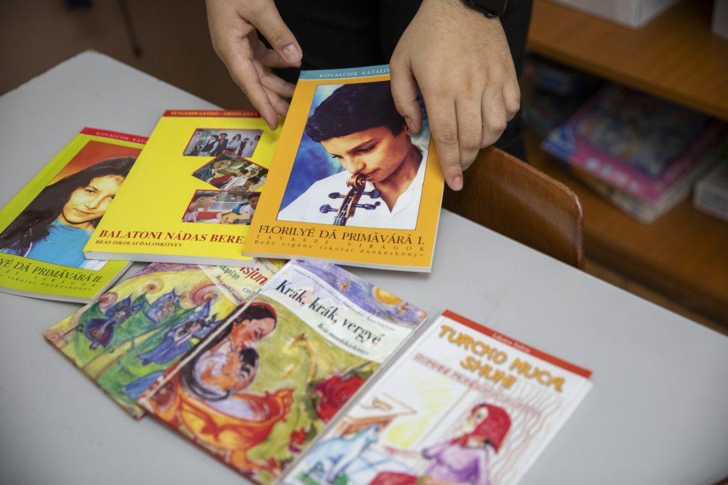 Egy kéz roma és beás nyelvű daloskönyveket és mesekönyveket válogat egy iskolai asztalon; a könyvek között gyermekeknek szóló kiadványok és kulturális tartalmak is szerepelnek.
